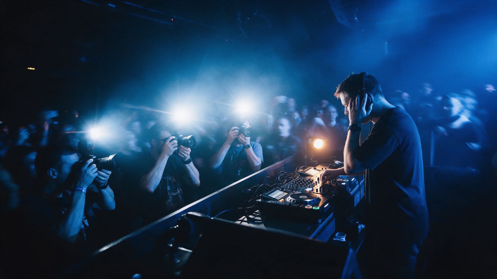 A DJ in a dark club booth, mid-mix, as photographers' flashes fire